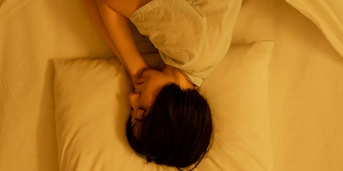 Top-down view of a person lying in bed under warm yellow light, resting with hands near the face and a pillow nearby.