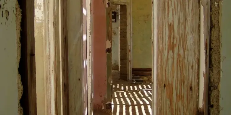 Sunlit corridor of an abandoned building with peeling walls and empty doorways, casting striped shadows on the dusty floor.
