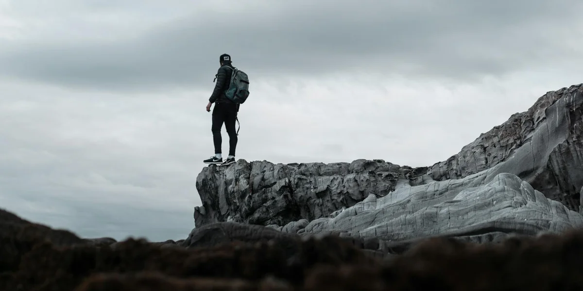 A lone hiker stands on the edge of a rocky cliff under a gray, overcast sky, gazing into the distance.