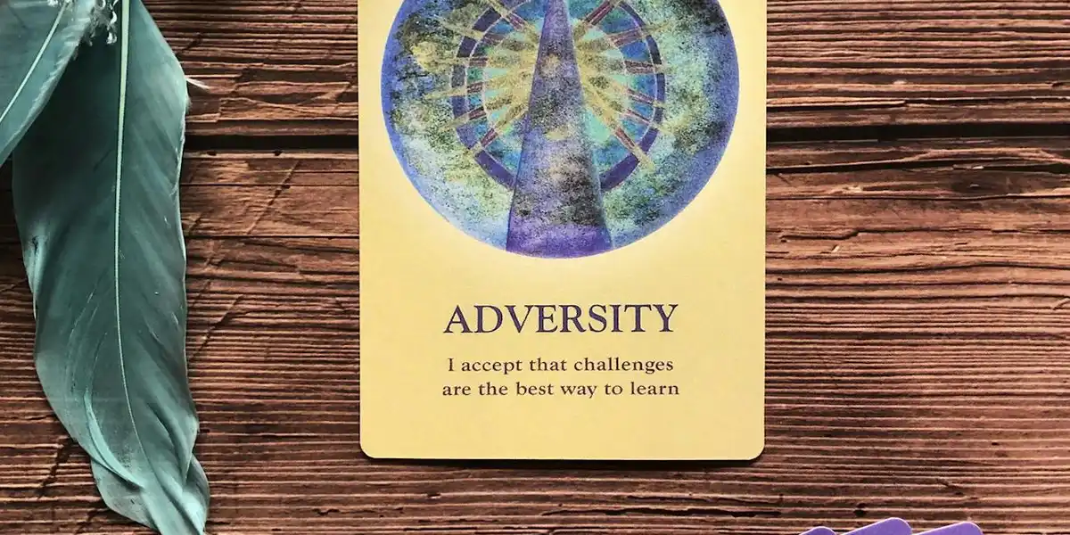 Yellow oracle card labeled 'ADVERSITY' with the subtitle 'I accept that challenges are the best way to learn', placed on a wooden surface beside a teal feather.