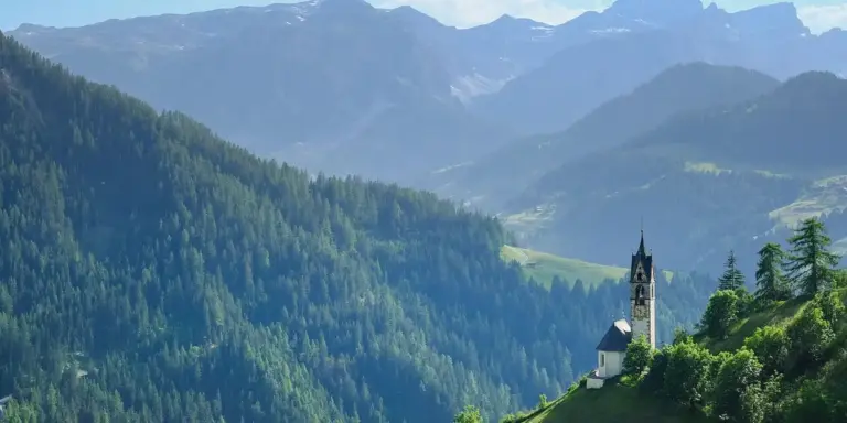 A small white church with a tall steeple sits on a hillside among dense evergreen trees, with blue, forested mountains in the distance.