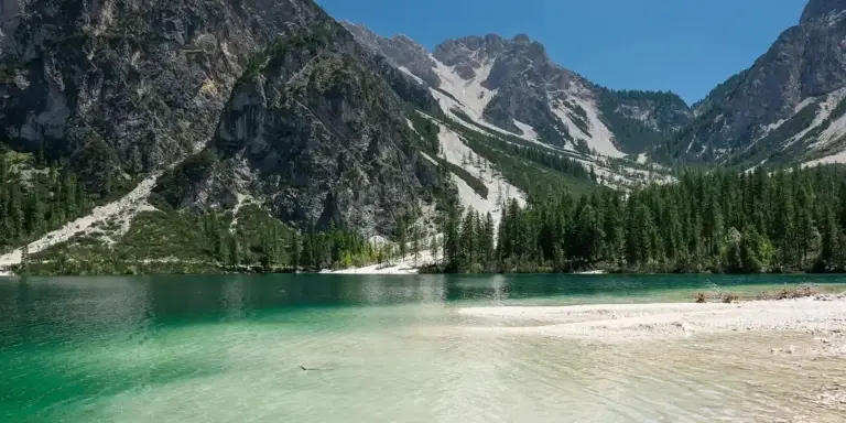 Turquoise alpine lake surrounded by evergreen trees and towering, snow-speckled mountains under a clear blue sky.