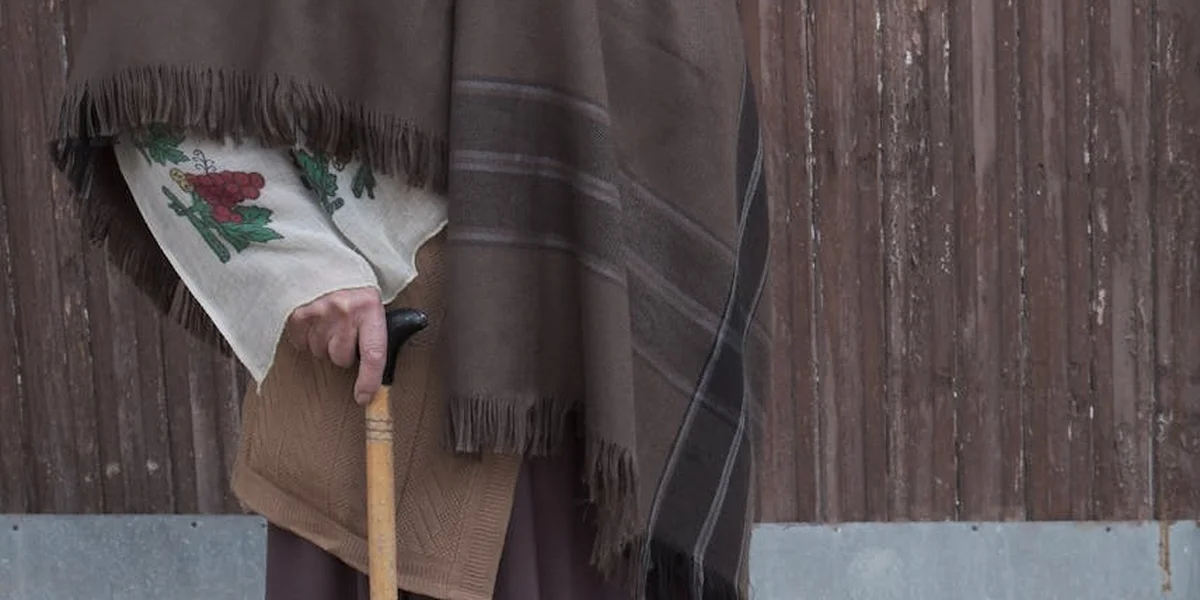 Close-up of a person wearing a fringed brown shawl over a patterned sleeve, gripping a wooden walking stick beside a weathered wooden fence.