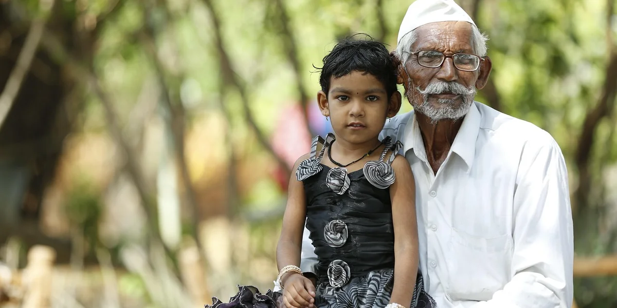 An elderly man in a white shirt and turban sits beside a young girl in a black dress in a sunlit park, symbolizing ancestral presence in dreams.