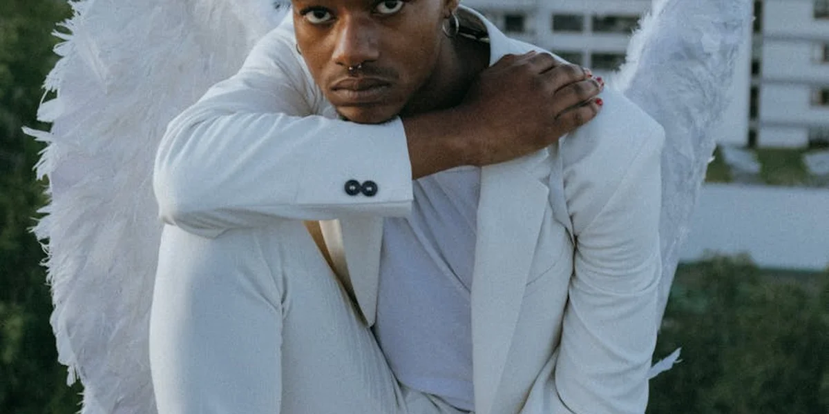 Man in a white suit with feathered angel wings outdoors, leaning forward with arms crossed.