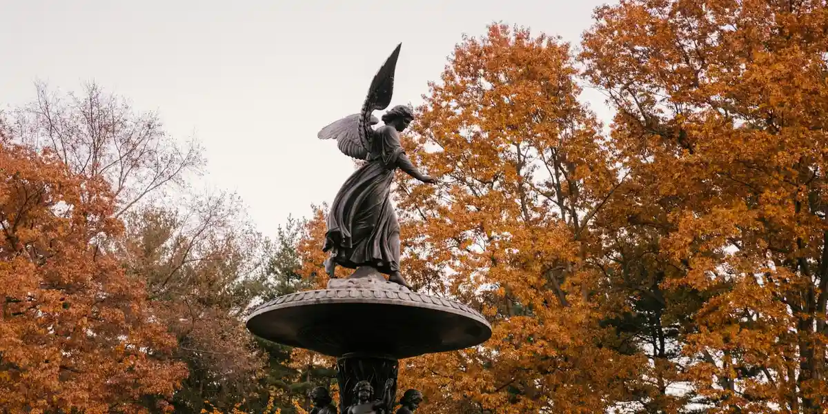 Statue of an angel perched on a fountain in a park with orange autumn trees