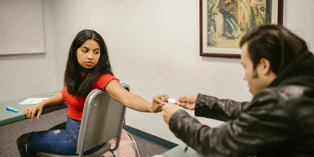 A young woman with dark hair wearing a red top sits at a table, looking anxious, as a man in a dark jacket offers something to her in a tense interaction inside a small room.