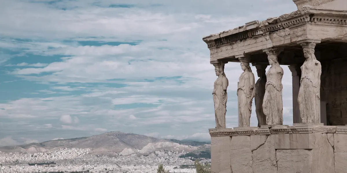 Ancient Greek temple facade with Caryatids (female statues) standing as columns, under a blue sky with distant mountains.