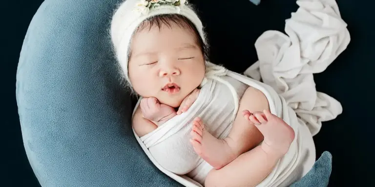 Newborn baby sleeping peacefully, wrapped in a white swaddle and wearing a delicate headband, resting on a blue cushion.