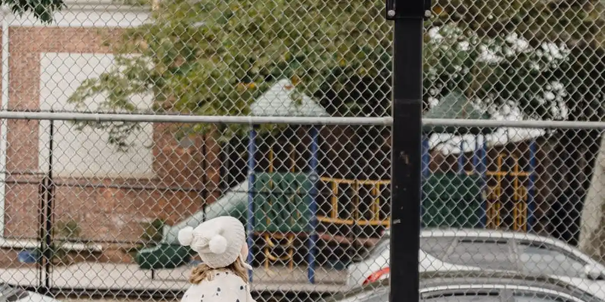 A person wearing a white hoodie stands near a chain-link fence with a playground and parked cars in the background.