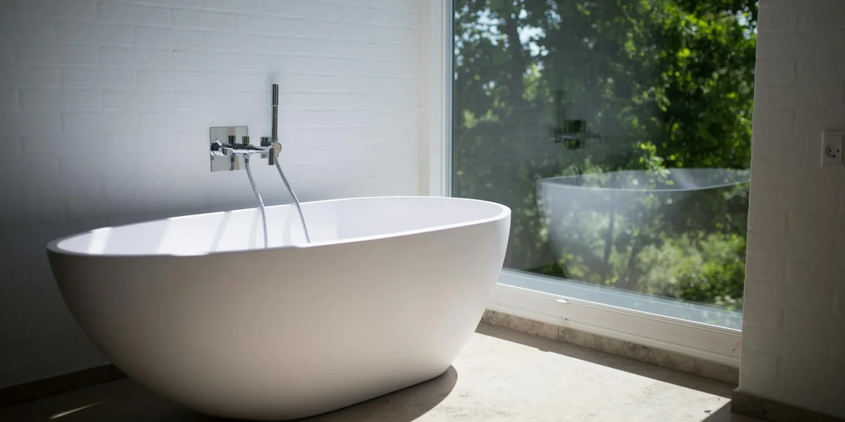 A clean, modern bathroom with a white freestanding bathtub beside a large window, letting in natural light and a view of greenery.