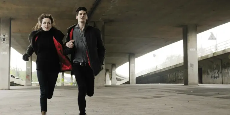 Two people run under a concrete overpass, looking tense as if they are being chased.