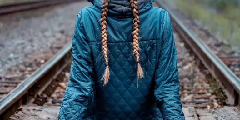 Person with two long braids wearing a blue quilted jacket, seen from behind, standing on railroad tracks.