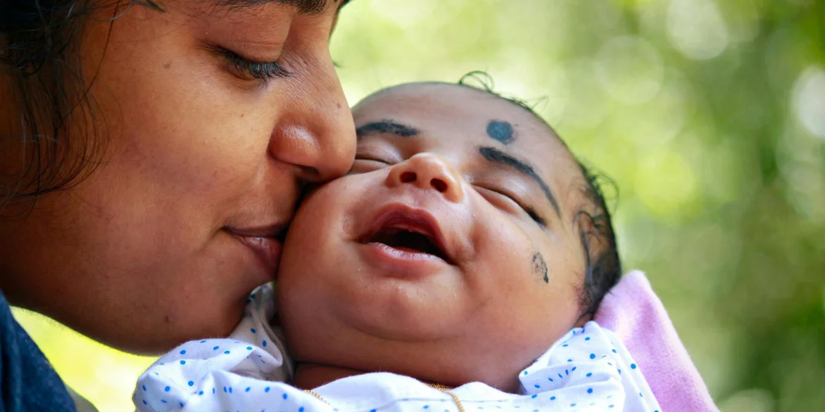 Close-up of a mother kissing a smiling newborn baby outdoors, symbolizing birth and nurturing.