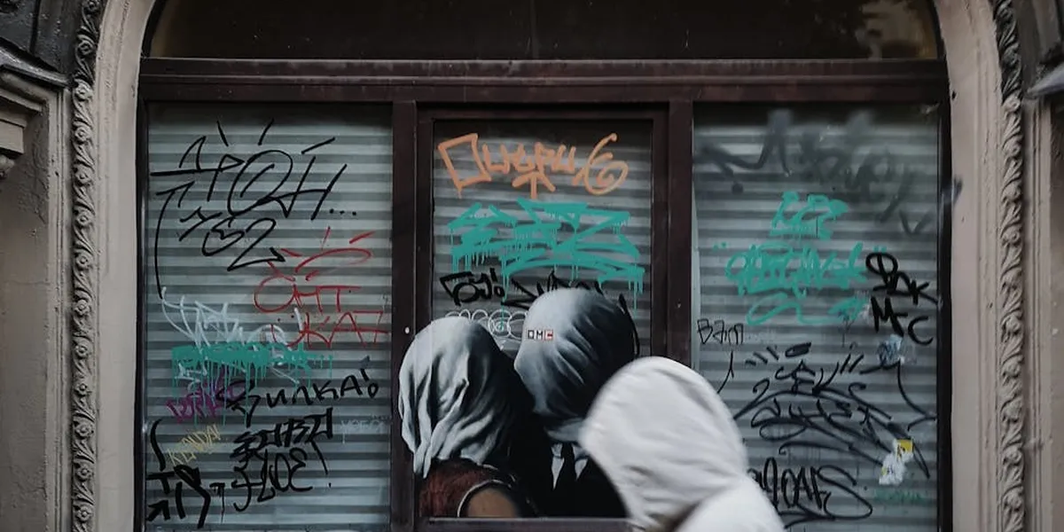Two hooded figures in white and gray cloaks stand in front of a graffiti-covered window, evoking mystery and dream symbolism.