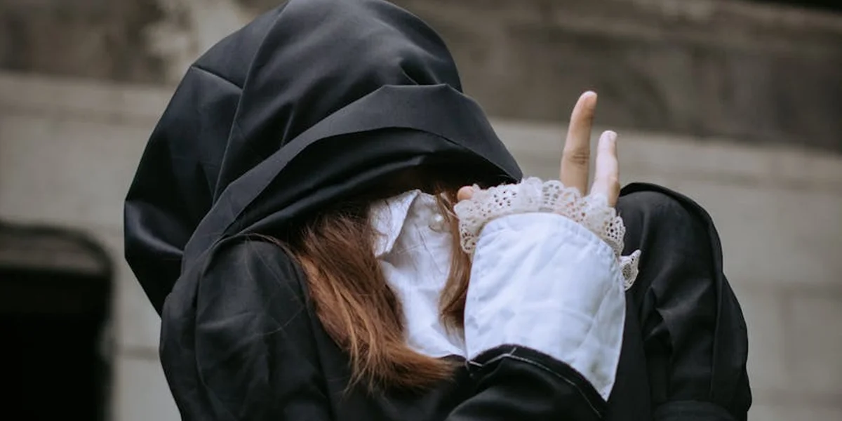 Close-up of a person wearing a dark hooded cloak with white lace cuffs, hands raised near the face against a stone background.