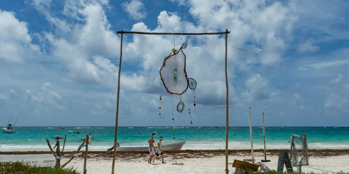 On a sunny beach, a simple wooden frame stands in the sand with a hanging shell-like sculpture and strings of beads, framed by the turquoise sea and blue sky.