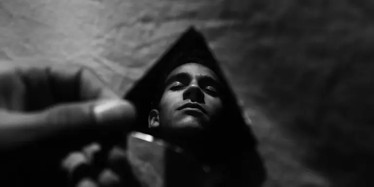 Grayscale portrait of a person looking upward, with a hand reaching from the left in the foreground and a dark triangular backdrop, creating a dreamlike, introspective mood about body and appearance.