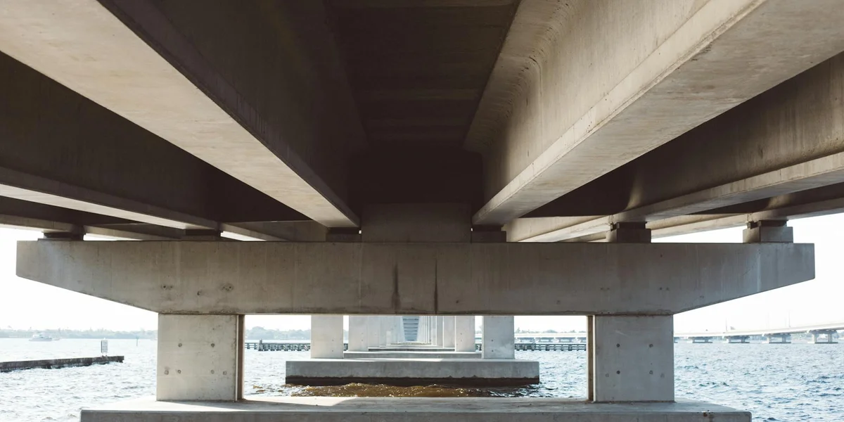 Underneath a concrete bridge over a body of water, showing large supports and crossing beams.