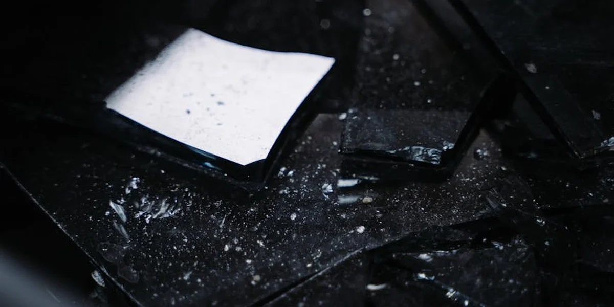 Close-up of broken glass shards scattered on a dark surface, with a small white note among the fragments.