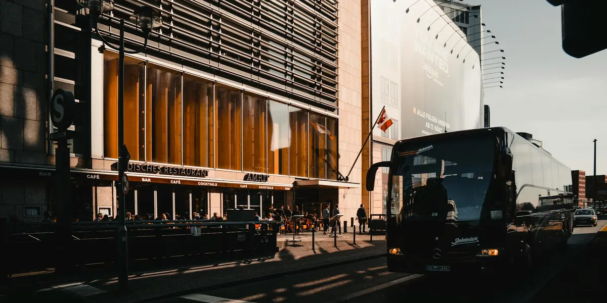 A large coach-style bus is parked at a curb on a city street, with a modern building and orange-lit storefront windows in the background and pedestrians nearby.