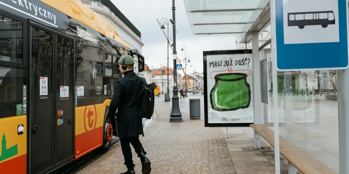 A person in a dark coat with a backpack stands beside a yellow city bus at a bus stop, with a glass shelter and a poster nearby.