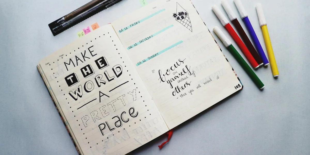 Open notebook on a light surface with the handwritten phrase 'MAKE THE WORLD A BETTER PLACE' and several colored markers nearby.