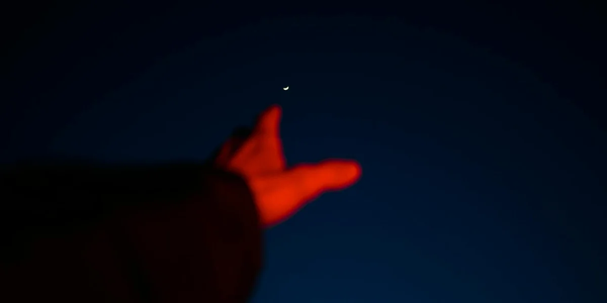 A hand in a dark red sleeve reaches toward a small bright light in a deep blue night sky.