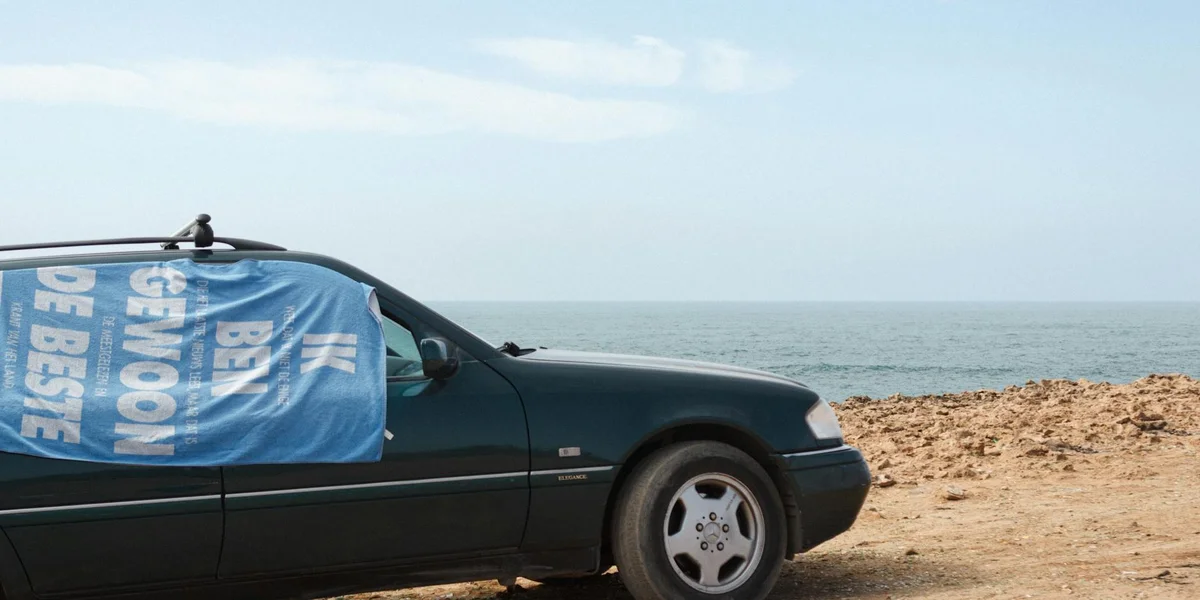 A dark car on a sandy beach with a blue banner draped over the side window, suggesting disruption.