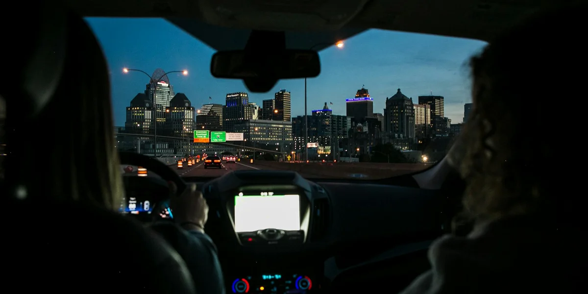 Interior view of a car at night with a city skyline lit up ahead, as passengers navigate a highway.