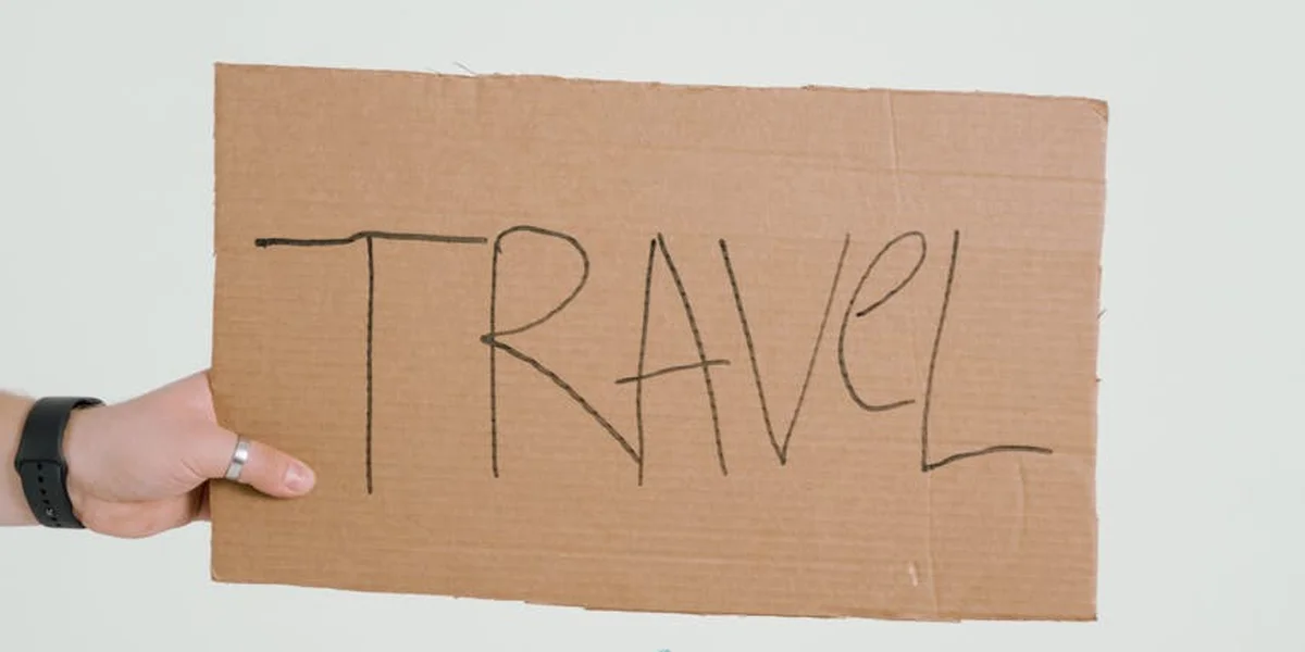 A hand wearing a black wristband holds a rectangular brown cardboard sign with the word 'TRAVEL' written in black marker, against a plain light background.