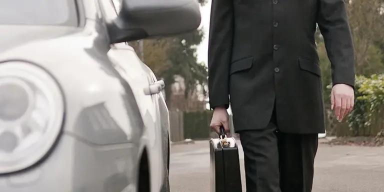 Businessperson in a black suit holding a briefcase, standing beside a parked car in a parking lot