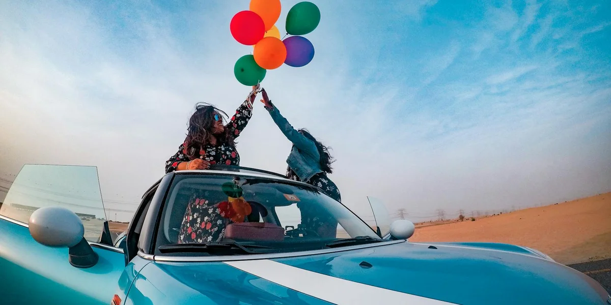 Two people in a blue convertible celebrate in the desert, raising colorful balloons from the open roof.