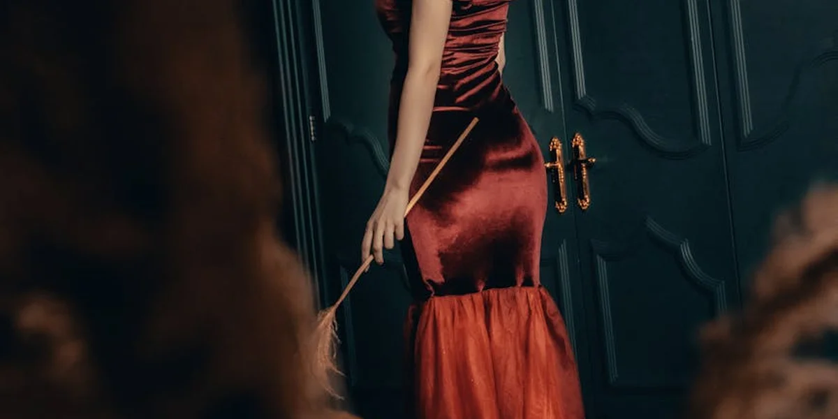 A person in a red satin gown stands with their back turned by a dark doorway, hinting at mystery and allure.