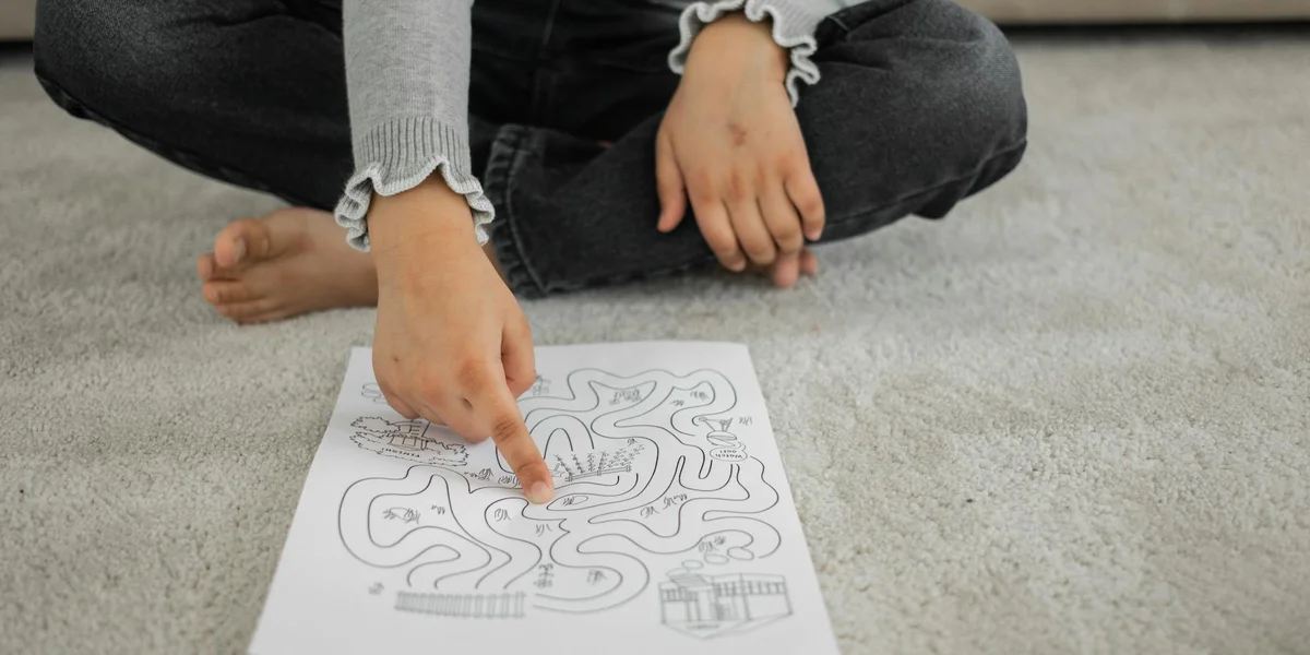 A child sits on a carpet, pointing at a maze drawing on a sheet of paper