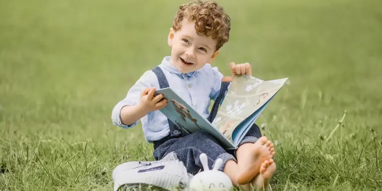 A joyful young child sitting on green grass, barefoot, reading a picture book outdoors