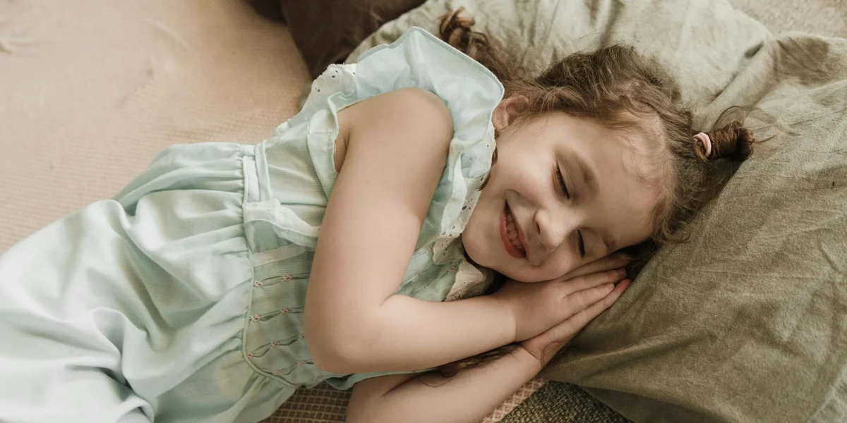 Young girl sleeping peacefully on a blanket, smiling softly with hands tucked under her cheek.
