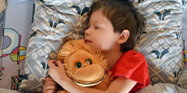 A young child sleeps in bed, cuddling a plush monkey.