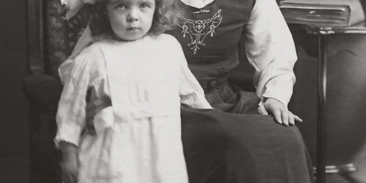 A black-and-white photograph of a young child seated beside an adult woman in a vintage dress, conveying a quiet, intimate mother-child moment.