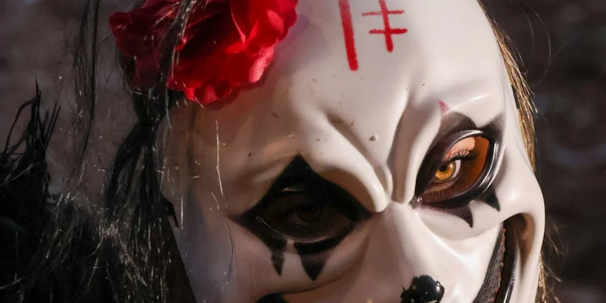 Close-up of a white clown mask with black eye makeup, orange eyes, a red flower, and a red symbolic mark on the forehead.