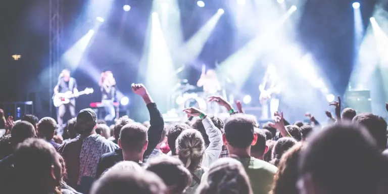 Crowd at a live concert, hands raised, with a band performing on a brightly lit stage.