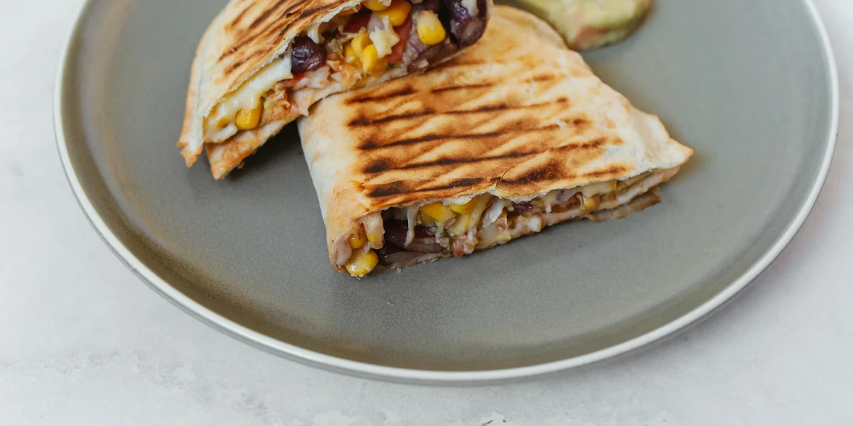 Grilled tortilla wrap cut in half on a gray plate, filled with corn kernels and vegetables.
