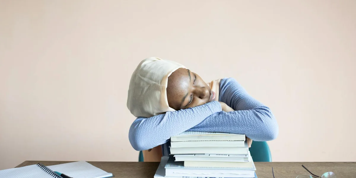 A person wearing a light hoodie rests their head on a stack of books on a desk, illustrating reliance on multiple references for dream symbolism.