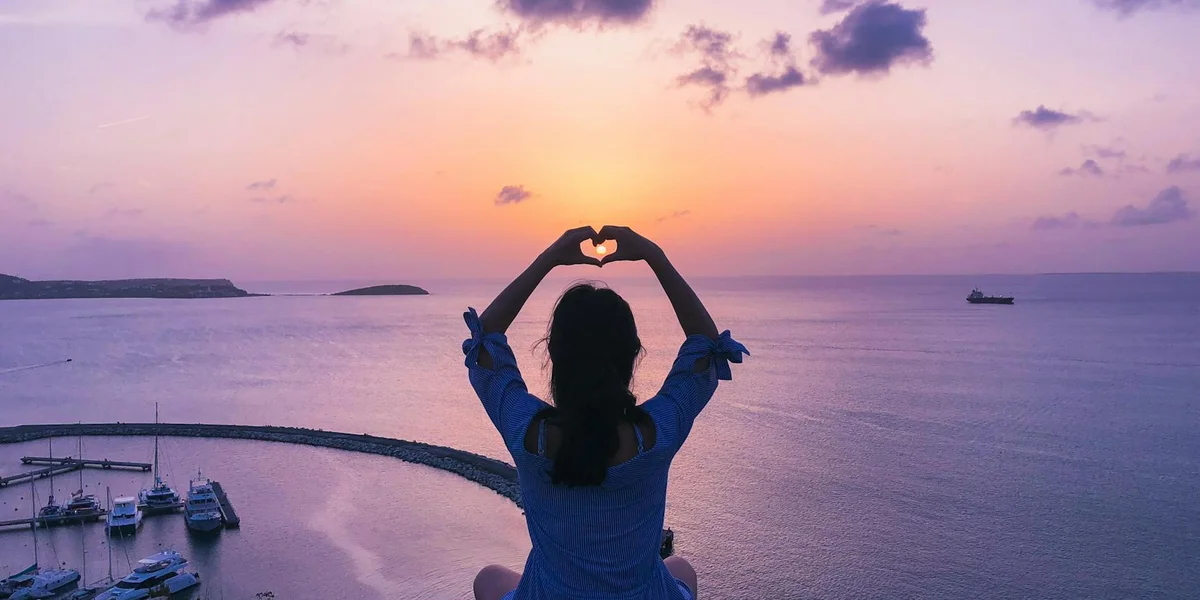 Silhouette of a person standing at the edge of the sea at sunset, arms raised above the head forming a heart shape