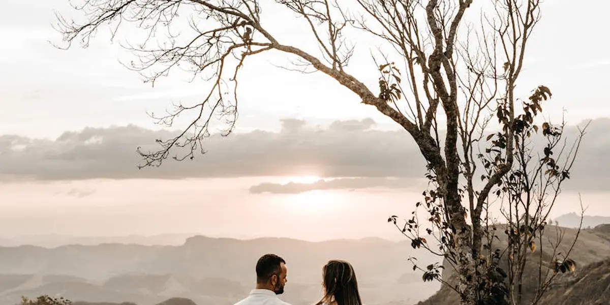 A silhouette of a couple sitting on a cliff edge at sunset, facing each other with a bare tree on the right and distant layered mountains under a soft, warm sky.