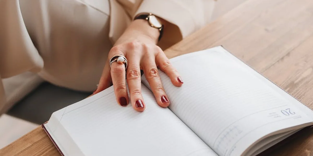 Close-up of a person’s hand with rings resting on an open notebook on a wooden surface.