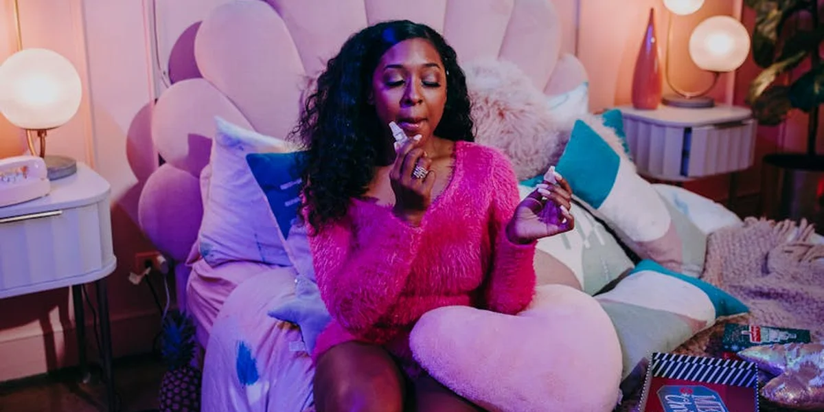 A person with dark curly hair wearing a bright pink sweater sits on a pastel bed surrounded by plush pillows in a softly lit, whimsical bedroom.
