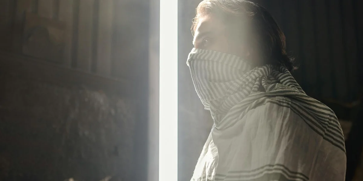 Profile of a person with a striped scarf covering their lower face, standing in a doorway with strong backlight.