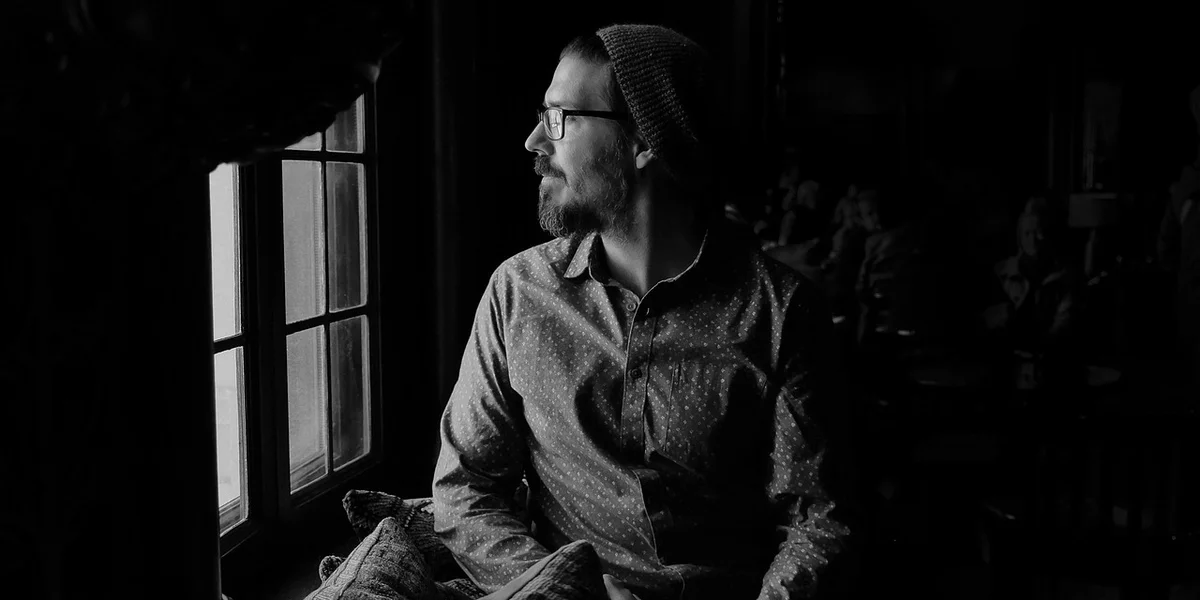 A man wearing a beanie and glasses sits in a dim, shadowy room, looking toward a bright window, symbolizing introspection and the spiritual meaning of darkness in dreams.