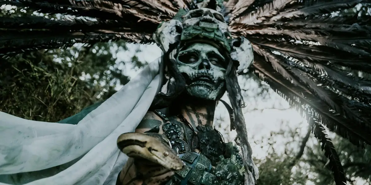 A skull-faced figure wearing a feathered headdress stands in a forest, evoking sudden danger and the symbolic strike of a snake.